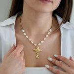 Collier doré avec perles blanches baroques et pendentif croix martelée sur une femme en chemise blanche.