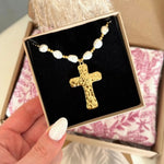 Collier croix martelée dorée, perles baroques blanches et perles dorées, dans un coffret velours noir, tenu par une main.