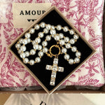 Collier de perles blanches et dorées avec croix en cristaux rectangulaires, dans boîte noire, sur tissu floral rose, étiquette AMOUR.