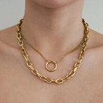 Colliers dorés superposés sur le cou. Un collier chaîne épaisse et un collier chaîne fine avec pendentif anneau.