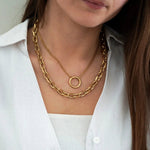 Femme portant deux colliers en or superposés: une chaîne épaisse à maillons et un collier fin avec pendentif anneau, sur une chemise blanche.