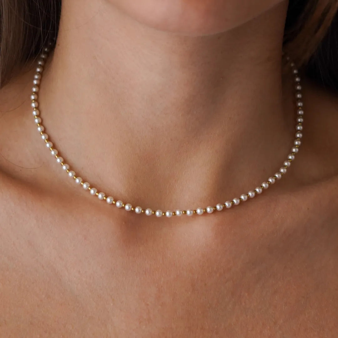 Collier ras-de-cou en perles blanches et billes d'acier inoxydable doré, porté sur le décolleté.