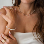 Collier ras-de-cou en perles blanches sur le cou d'une femme. Bracelet et bagues en acier inoxydable doré sont visibles.