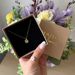 Collier fin en acier inoxydable doré, avec un pendentif fleur délicat, présenté dans son écrin marron tenu par une main.