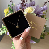 Collier fin en acier inoxydable doré, avec un pendentif fleur délicat, présenté dans son écrin marron tenu par une main.