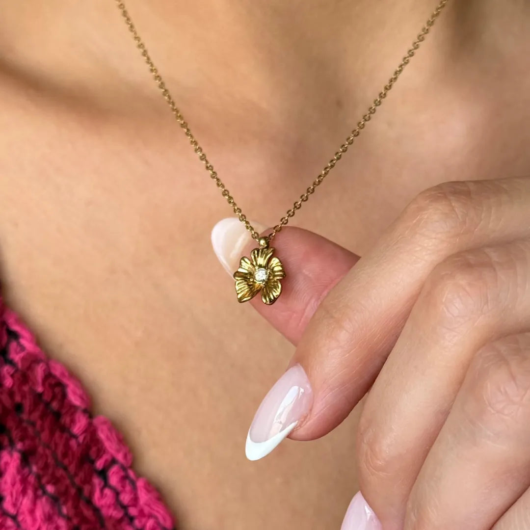 Collier à pendentif fleur en acier inoxydable doré, avec un brillant central, tenu par la main d'une femme sur son décolleté.