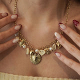 Gros plan sur le collier à breloques cœurs et perles MAISON DE L'AMOUR porté sur un top blanc côtelé, mise en valeur des détails dorés et des bagues.