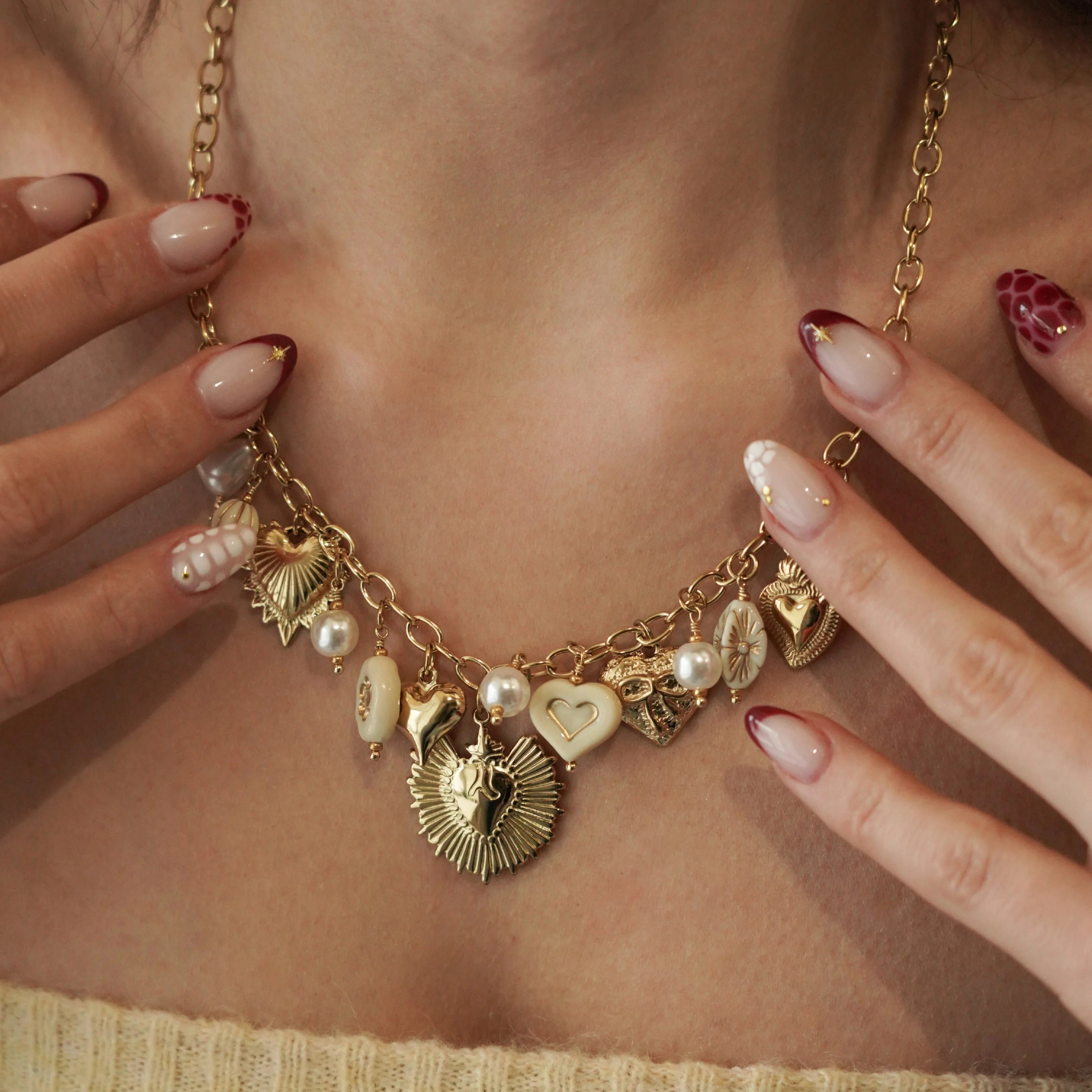 Gros plan sur le collier à breloques cœurs et perles MAISON DE L'AMOUR porté sur un top blanc côtelé, mise en valeur des détails dorés et des bagues.