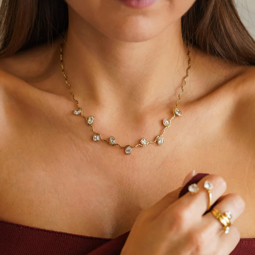 Collier vague en acier inoxydable doré, avec des zircons multiformes sertis clos, porté sur un décolleté féminin.