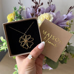 Collier acier inoxydable doré, pendentif fleur stylisé, présenté dans son écrin. Une main porte une bague fleur en nacre.