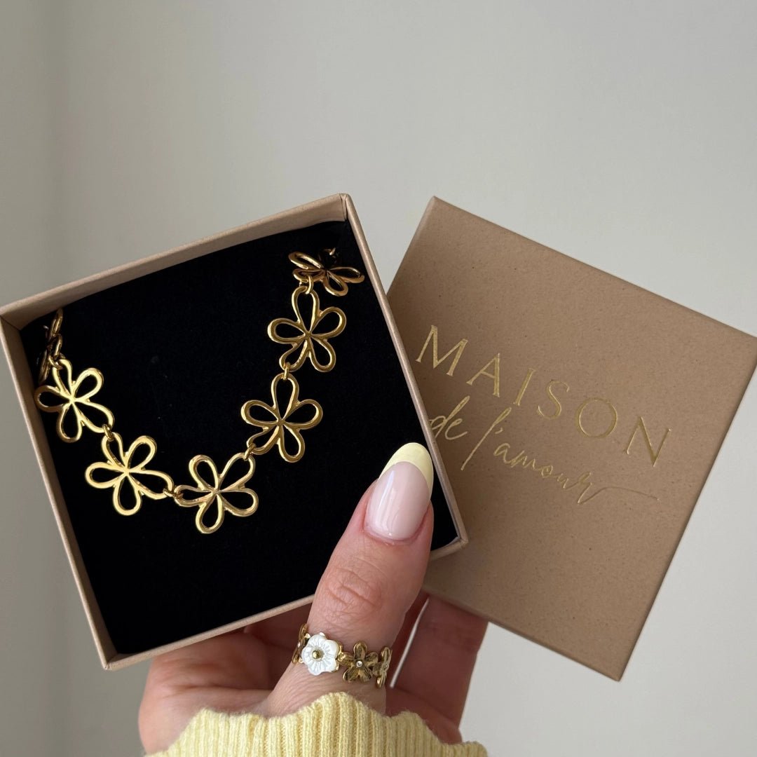 Collier acier inoxydable doré, design fleurs évidées, dans sa boîte cadeau "MAISON de l'amour". Main tenant l'écrin.