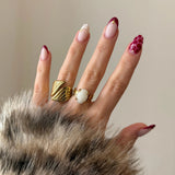 Duo de bagues dorées portées à l'index et au majeur, mises en valeur par une manucure graphique rouge.