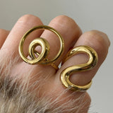 Accumulation de deux grosses bagues dorées sculpturales (modèle spirale et modèle vague) portées sur la même main.