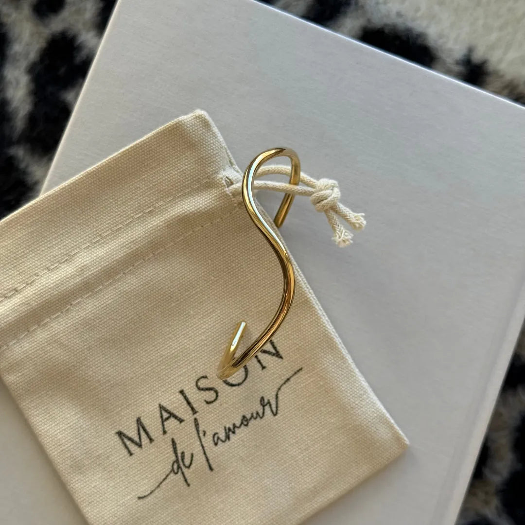Bracelet jonc ouvert en acier inoxydable doré à double vague asymétrique, sur un pochon en tissu "MAISON de l'amour" et une