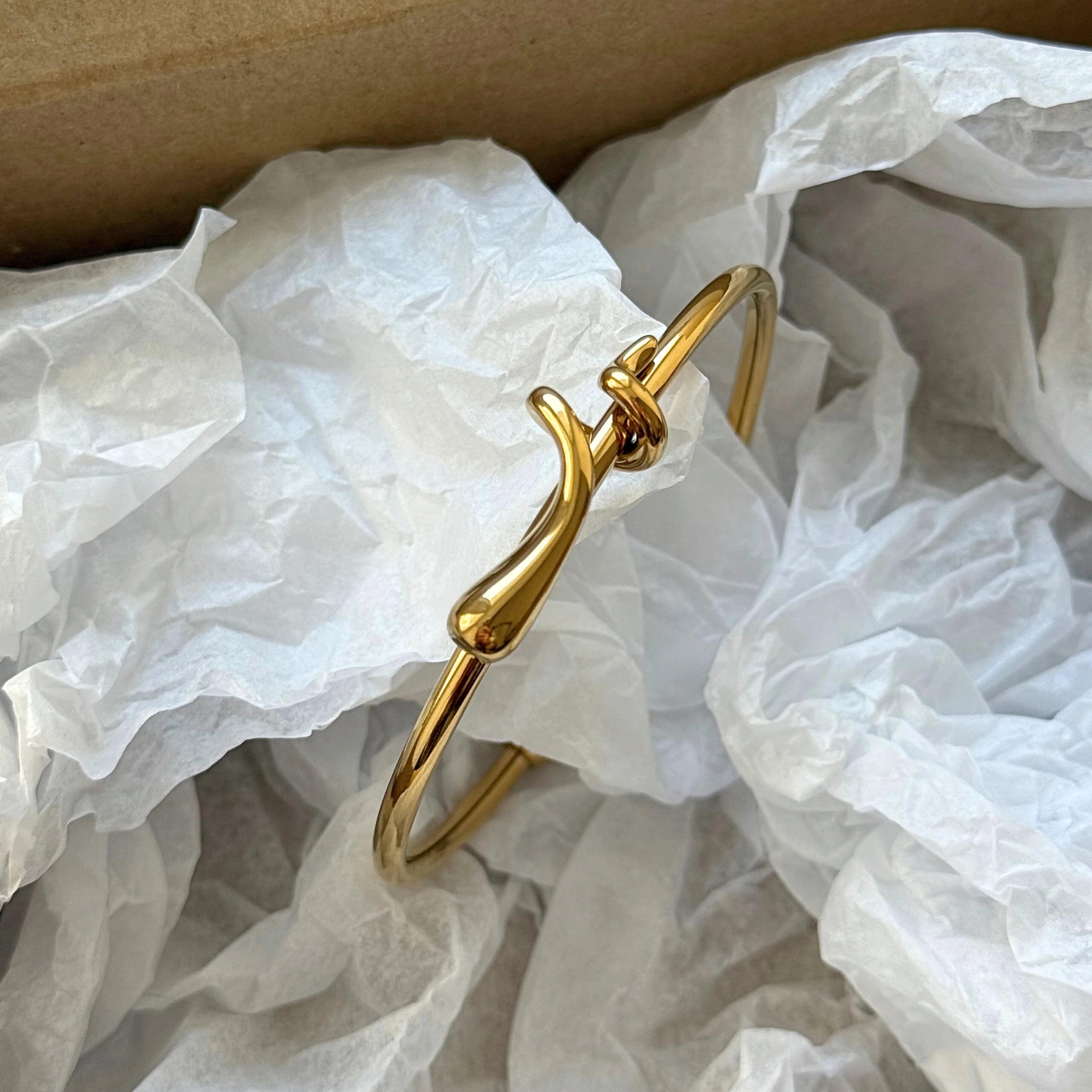 Bracelet jonc doré présenté dans son emballage d'expédition, niché dans du papier de soie blanc froissé.