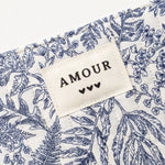 Pochette matelassée à motif floral bleu et blanc. Une étiquette beige cousue affiche "AMOUR" et trois cœurs noirs.