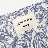 Pochette matelassée à motif floral bleu et blanc. Une étiquette beige cousue affiche "AMOUR" et trois cœurs noirs.