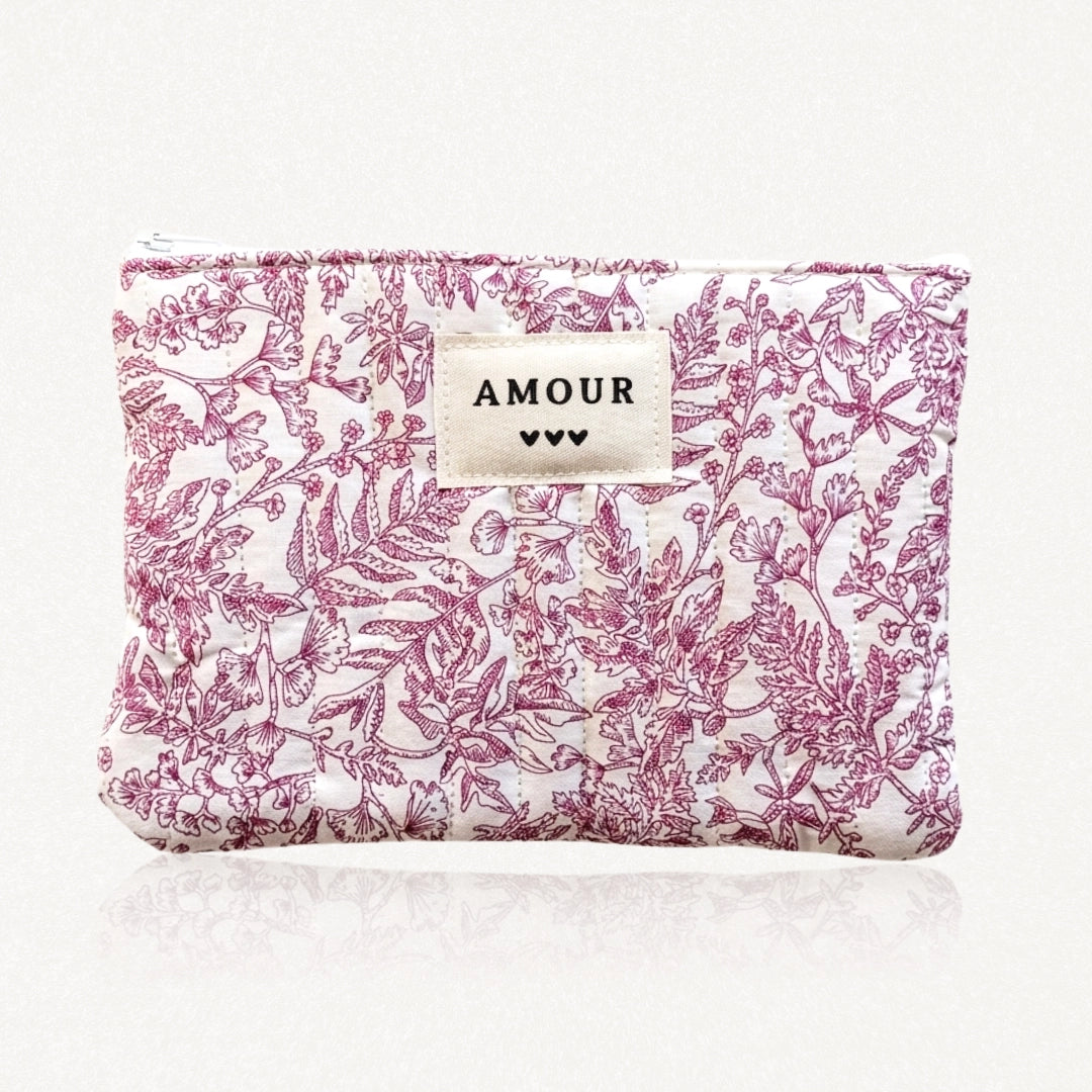 Pochette matelassée blanche à motif floral et feuillage magenta. Étiquette AMOUR avec trois cœurs noirs. Fermeture éclair