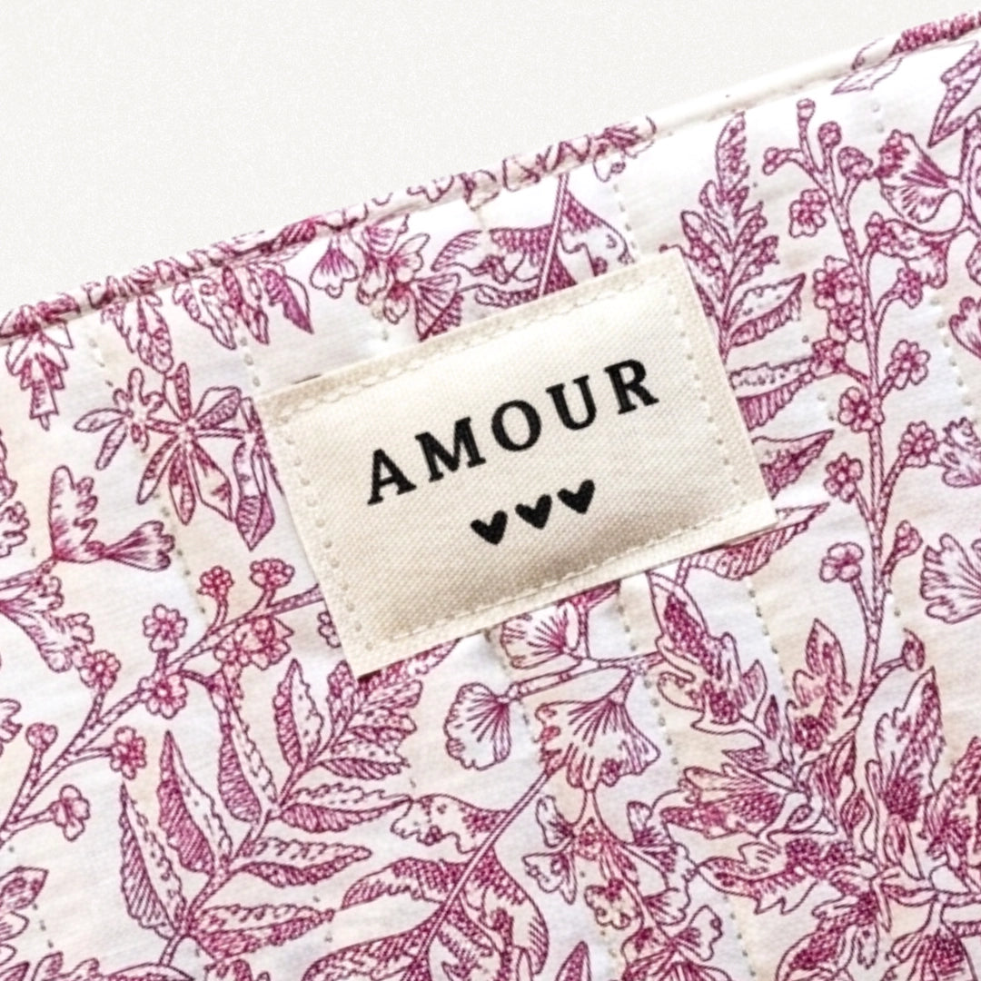 Gros plan sur une pochette matelassée blanche à motif floral rose, avec une étiquette "AMOUR" et trois cœurs.