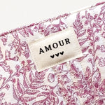 Gros plan sur une pochette matelassée blanche à motif floral rose, avec une étiquette "AMOUR" et trois cœurs.