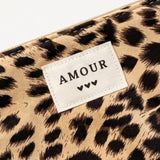 Pochette à motif léopard avec étiquette crème cousue "AMOUR" en majuscules noires et trois petits cœurs. Fermeture éclair