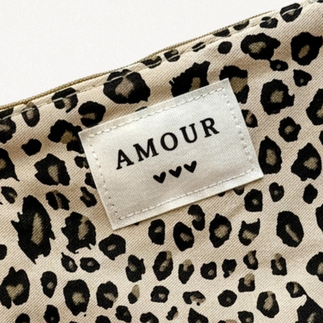 Gros plan d'une pochette beige à imprimé léopard. Une étiquette blanche cousue affiche "AMOUR" et trois cœurs noirs. Zip
