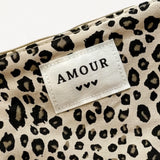 Gros plan d'une pochette beige à imprimé léopard. Une étiquette blanche cousue affiche "AMOUR" et trois cœurs noirs. Zip