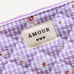 Pochette matelassée lilas à carreaux vichy avec marguerites blanches. Étiquette "AMOUR" et 3 cœurs noirs.