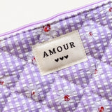 Pochette matelassée lilas à carreaux vichy avec marguerites blanches. Étiquette "AMOUR" et 3 cœurs noirs.