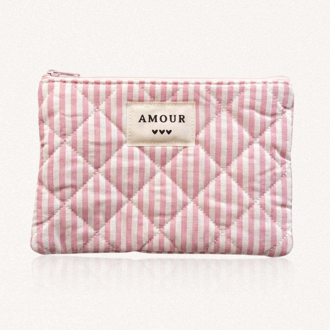 Pochette matelassée à rayures roses et blanches. Fermeture éclair rose. Étiquette crème "AMOUR" et 3 cœurs noirs.