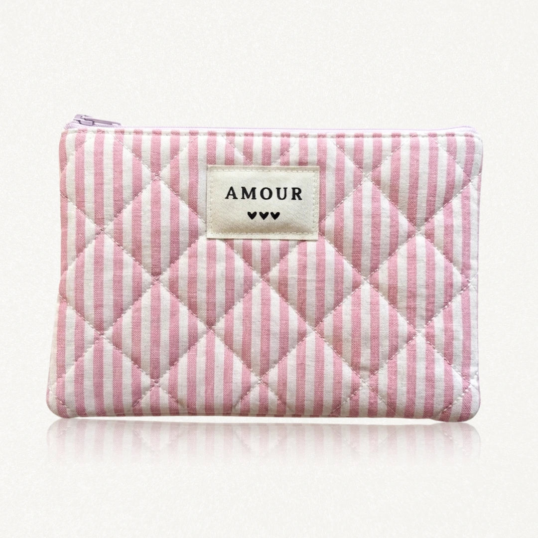 Pochette matelassée à rayures roses et blanches. Fermeture éclair rose. Étiquette crème "AMOUR" et 3 cœurs noirs.