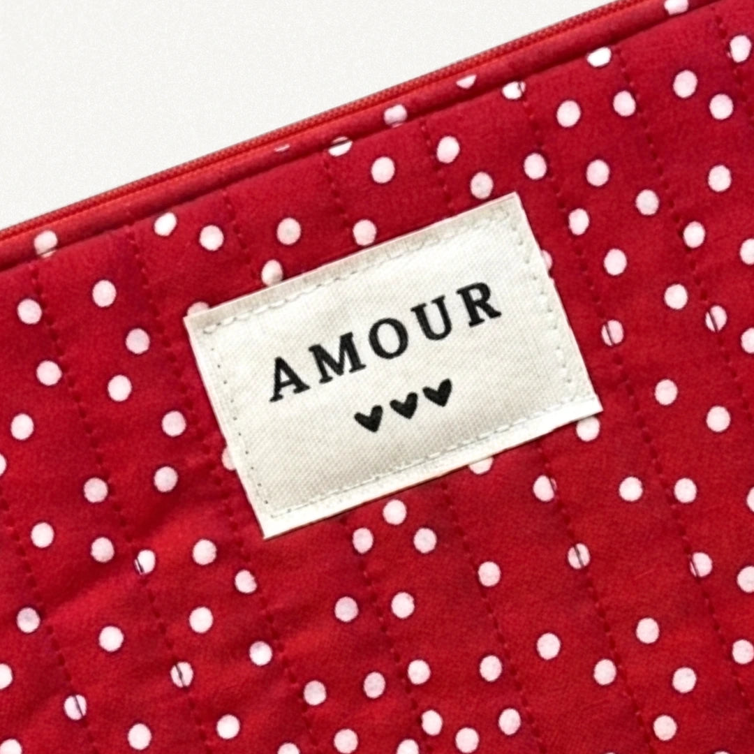 Pochette matelassée rouge à pois blancs. Étiquette beige brodée AMOUR et trois petits cœurs noirs. Fermeture éclair rouge.