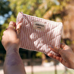 Pochette matelassée bijou en acier inoxydable rose rayé blanc, avec étiquette "AMOUR", tenue par des mains.