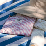 Pochette matelassée rose à rayures blanches avec l'inscription AMOUR, posée sur une serviette bleue près d'un chapeau de