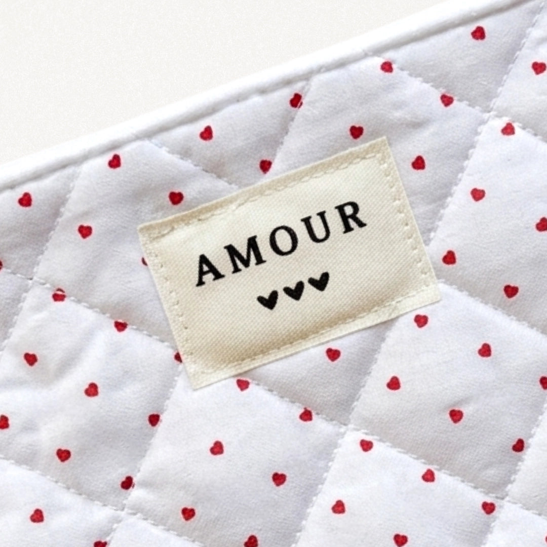 Pochette matelassée blanche à petits cœurs rouges, avec une étiquette "AMOUR" et trois cœurs noirs.