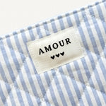 Pochette matelassée en tissu rayé blanc et bleu clair avec étiquette "AMOUR" et 3 cœurs noirs.