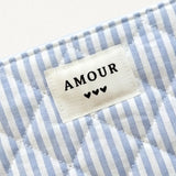 Pochette matelassée en tissu rayé blanc et bleu clair avec étiquette "AMOUR" et 3 cœurs noirs.