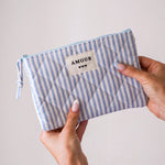 Pochette textile matelassée à rayures verticales blanches et bleues. Étiquette "AMOUR" et 3 cœurs noirs, tenue par deux