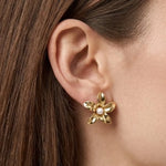 Boucle d'oreille puce fleur en acier inoxydable doré, avec une perle blanche au centre, portée sur une oreille.
