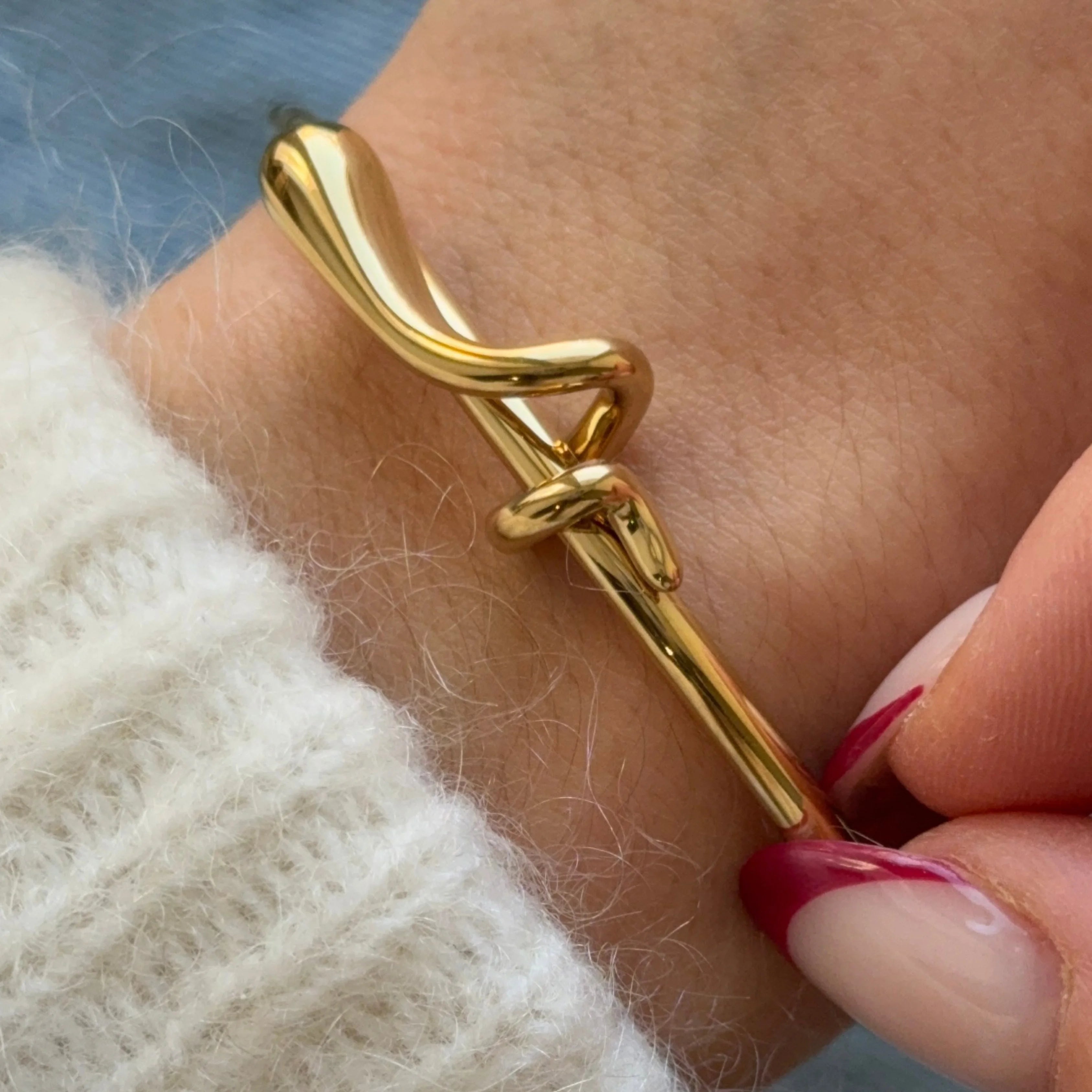 Bracelet jonc doré à nœud porté au poignet, associé à un pull en maille blanche douce pour un look hivernal.